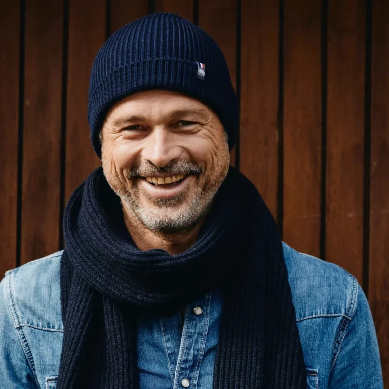 Personne debout devant un mur en bois, portant un bonnet bleu marine, une écharpe assortie et une chemise en jean, avec le visage flouté.